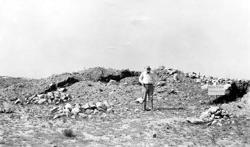 Unknown researcher poses in front of Ruin 55. Label reads: "Partly excavated pueblo ruin (digging by Studer, Mason, Moorehead, et al.) Photo by A.T. Jackson, 1935." Sign warns trespassers against disturbing investigations at site. TARL Archives