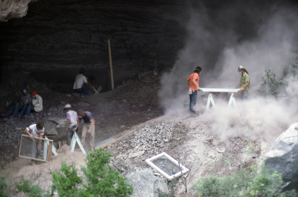 photograph of people excavating and screening dirt in a big desert rockshelter, stirring up a huge cloud of dust