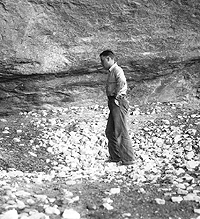 Man stands in "sotol pit" in Fate Bell Shelter, 1932. This large rock-strewn depression is a roasting pit where sotol, lechuguilla, and probably other plants were baked. There were several such pits visible in the shelter when the archeologists arrived. Photo from ANRA-NPS Archives at TARL.