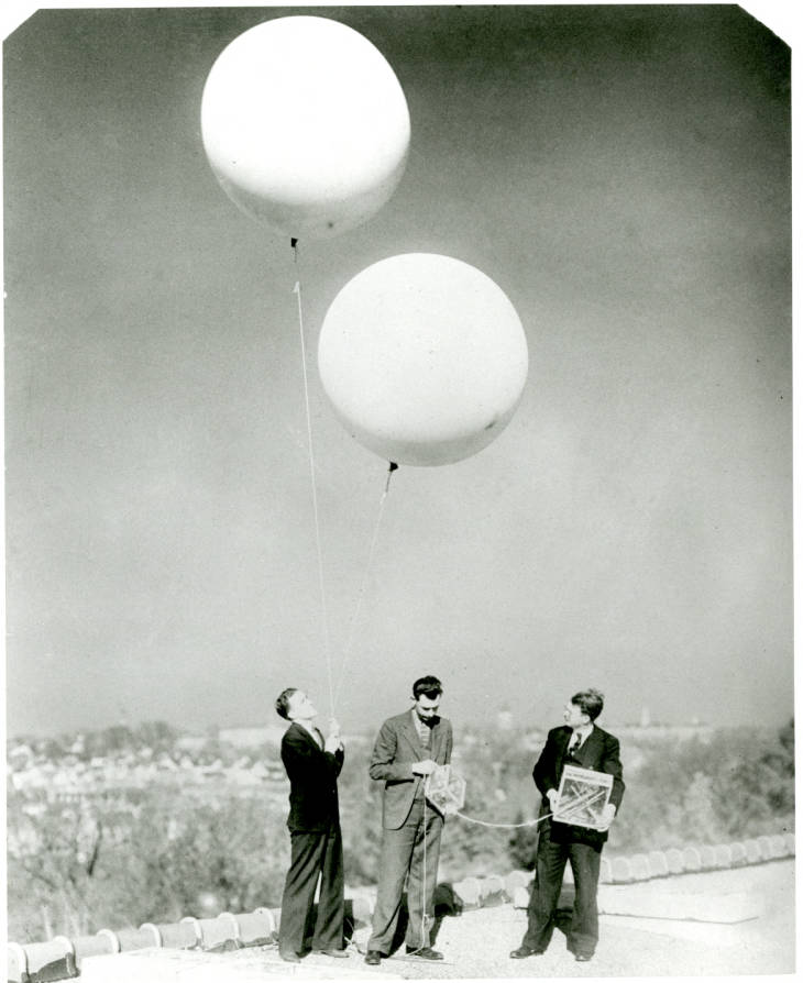Black and white photograph of three men on a hillside with two large pale balloons.