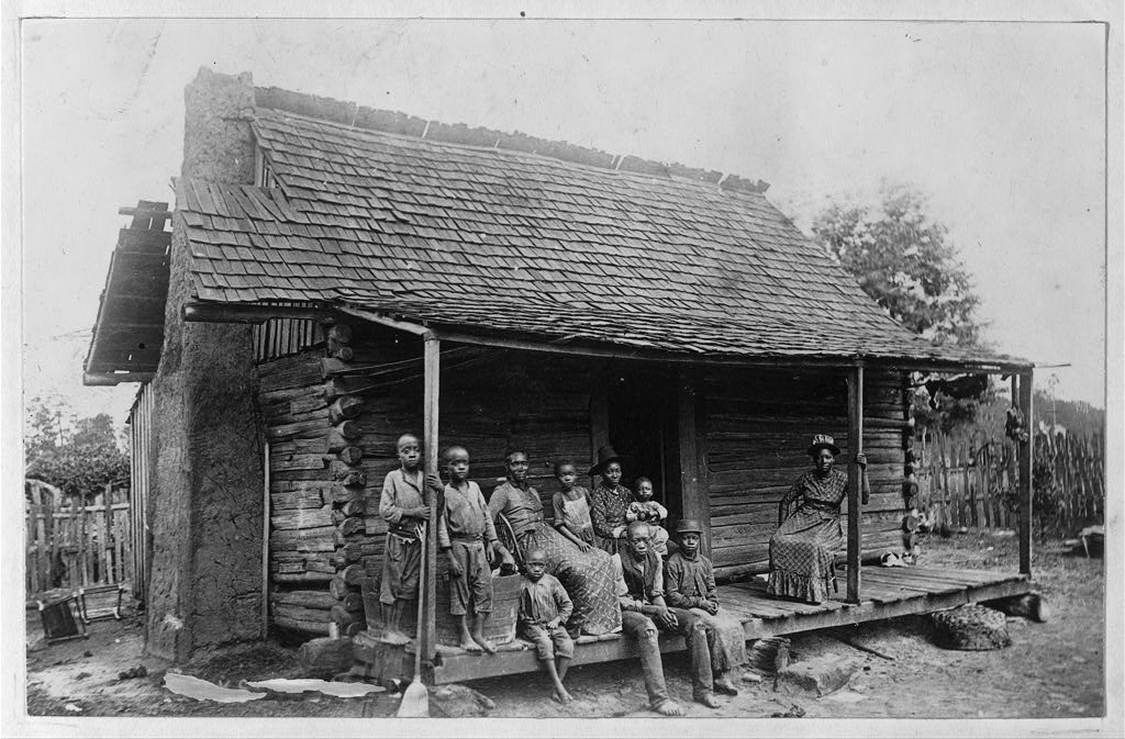 Black and white photograph of ten African Americans sitting on the porch of a log cabin with a brick chimney. A fence is in the background, and the area around the house is bare dirt.