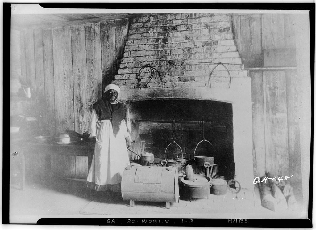 B&W photo the interior of a kitchen wall at the center of which is a fireplace in front of which a standing Black woman (cook) poses along with an array of cooking pots and pieces of equipment