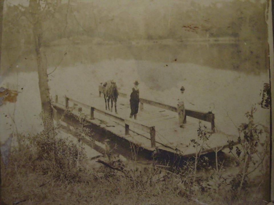 Sepia photograph of wooden river ferry on river bank, with two peole and a pack animal on board.