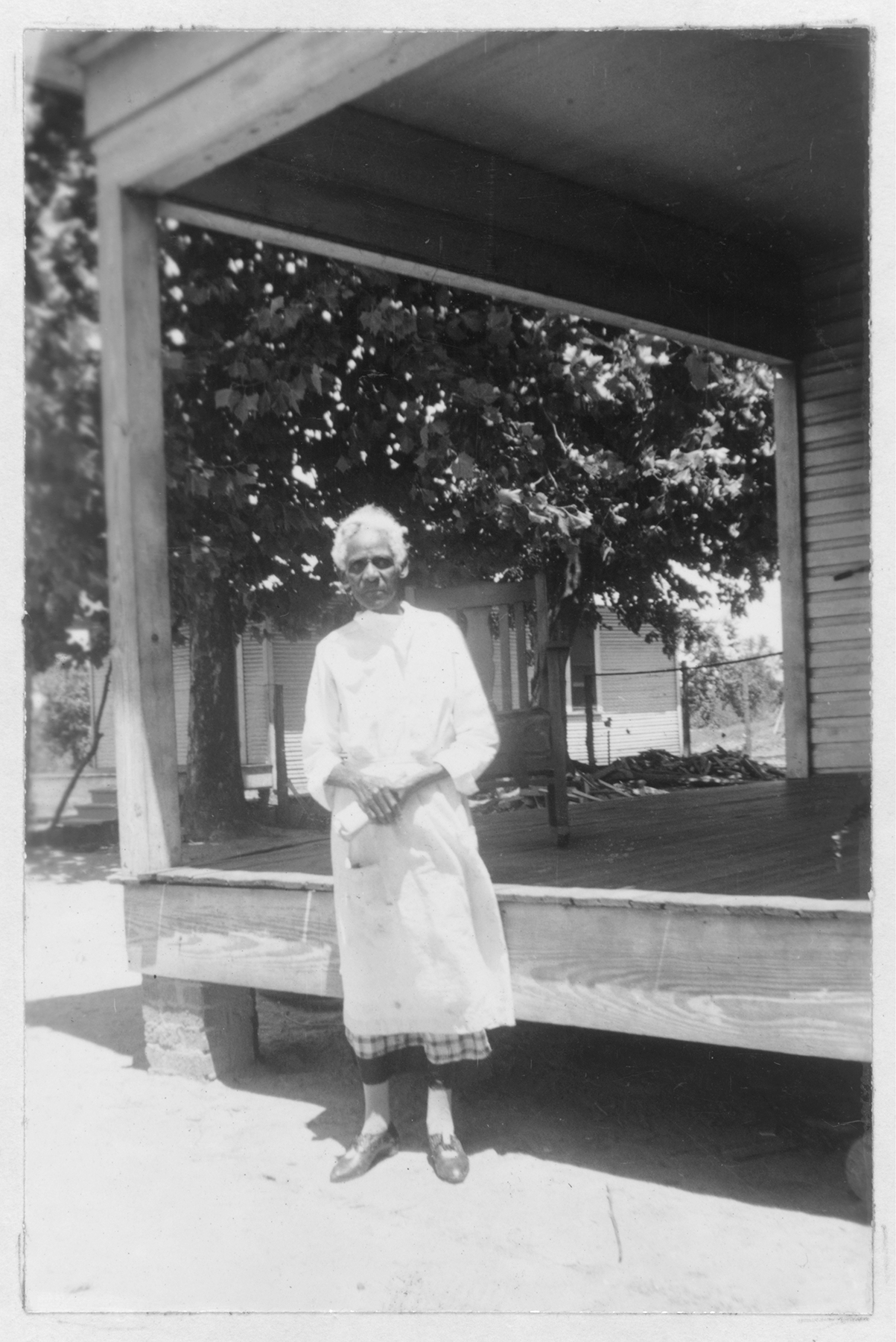 A black and white photograph of an older African American woman wearing a long white dress, standing in front of a house porch, upon which sits a wood rocking chair. A large tree is in the background, and it is a sunny day.