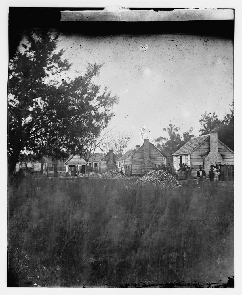 faded black and white photo of with cluster of sizeable log cabins in background seen through a tree on the left in foreground