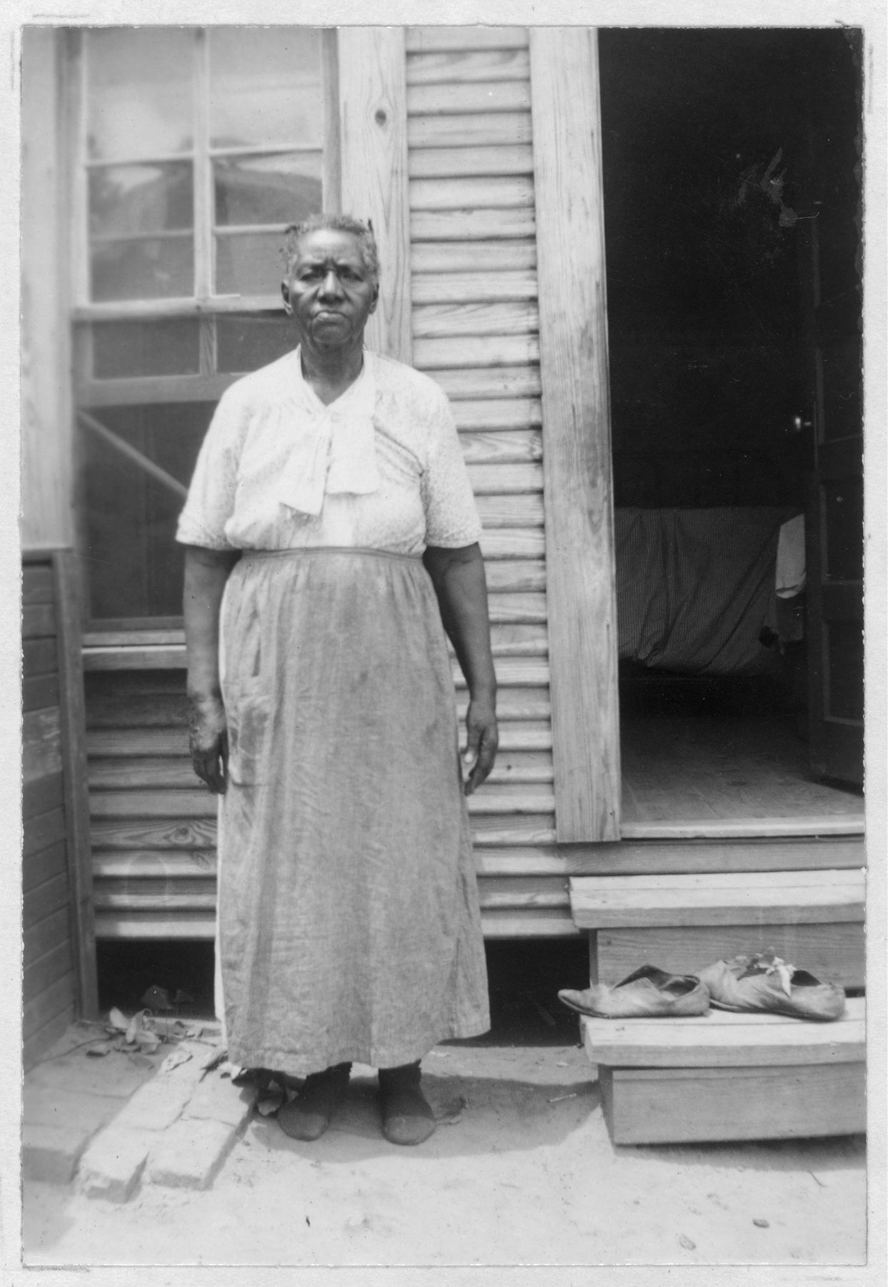 An older African American woman standing in front of a wood house. The woman wears a blouse and long skirt and socks. A pair of shoes are on the stoop on the right.