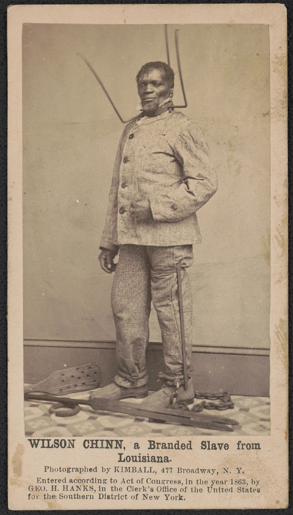 An enslaved African American man posing with equipment of torture in front of a wall.