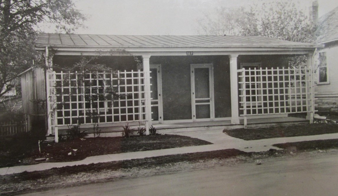black and white photograph of a house with a porch and trellises flanking the front door