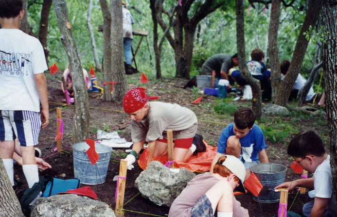 photograph of kids excavating archeology units in a forest