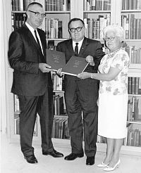 B#W photo of three formally dressed people standing in front of bookshelf.