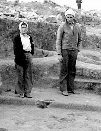 B&W photo of two young adults bundled up for cool weather and standing in excavated Antelope Creek house.