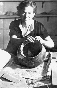 B&W photo of seated young woman facing camera; she is working on (cleaning) a large earthenware pottery jar.