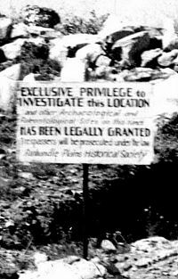 B&W photo of sign on edge of trench.