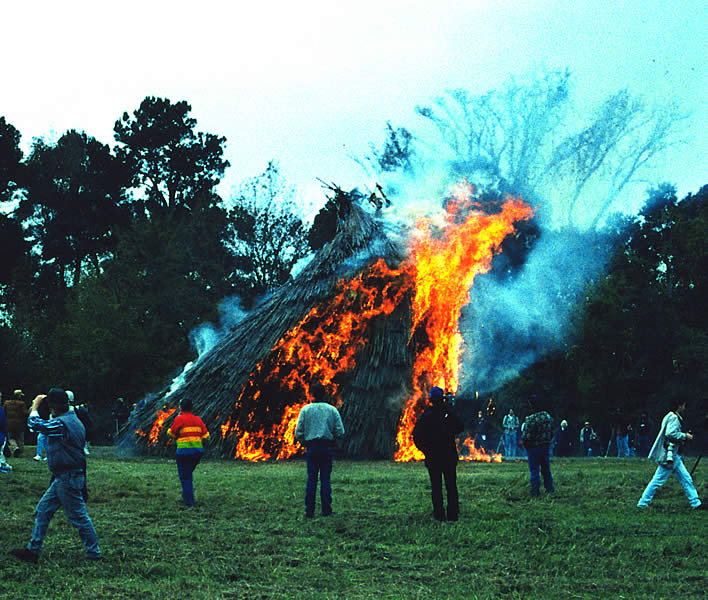 a grass house about half engulfed in big orange flames, as people look on