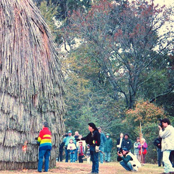 a crowd of people standing to the side of a grass house looking on as someone lights the house on fire