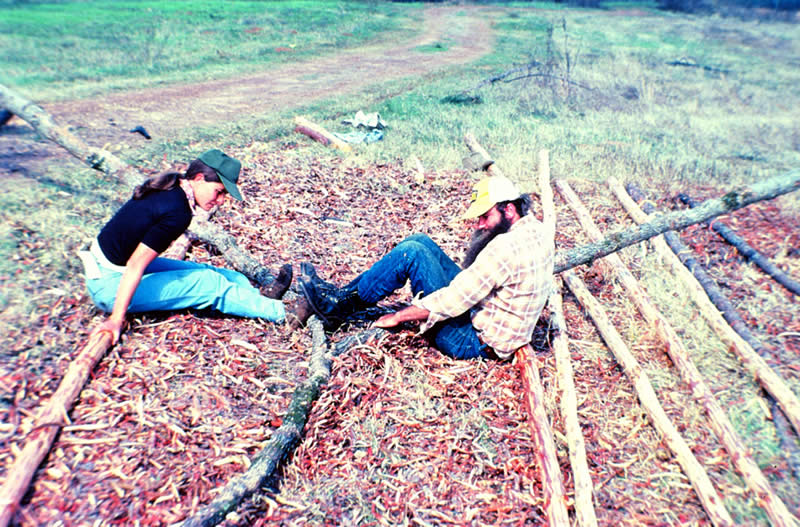 photograph of two people sitting on the ground with wood poles around them, bending one