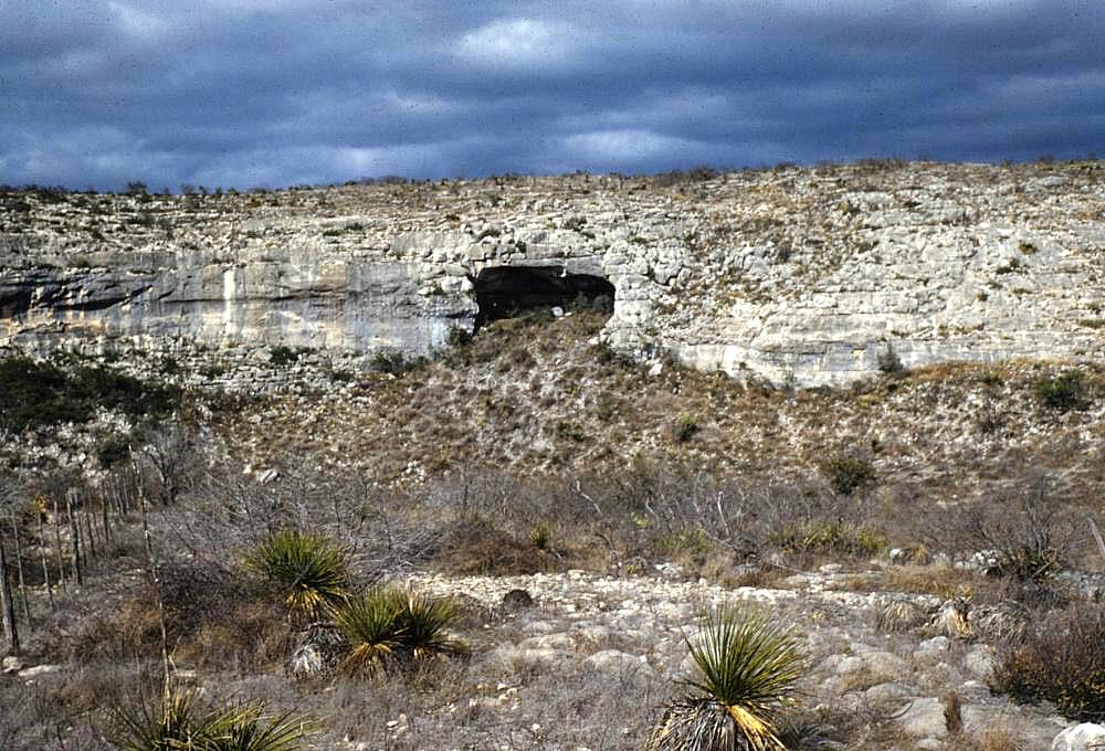 Photograph of a rock bluff with a large dark shelter entrance in its face.