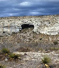 Photograph of a rock bluff with a large dark shelter entrance in its face.