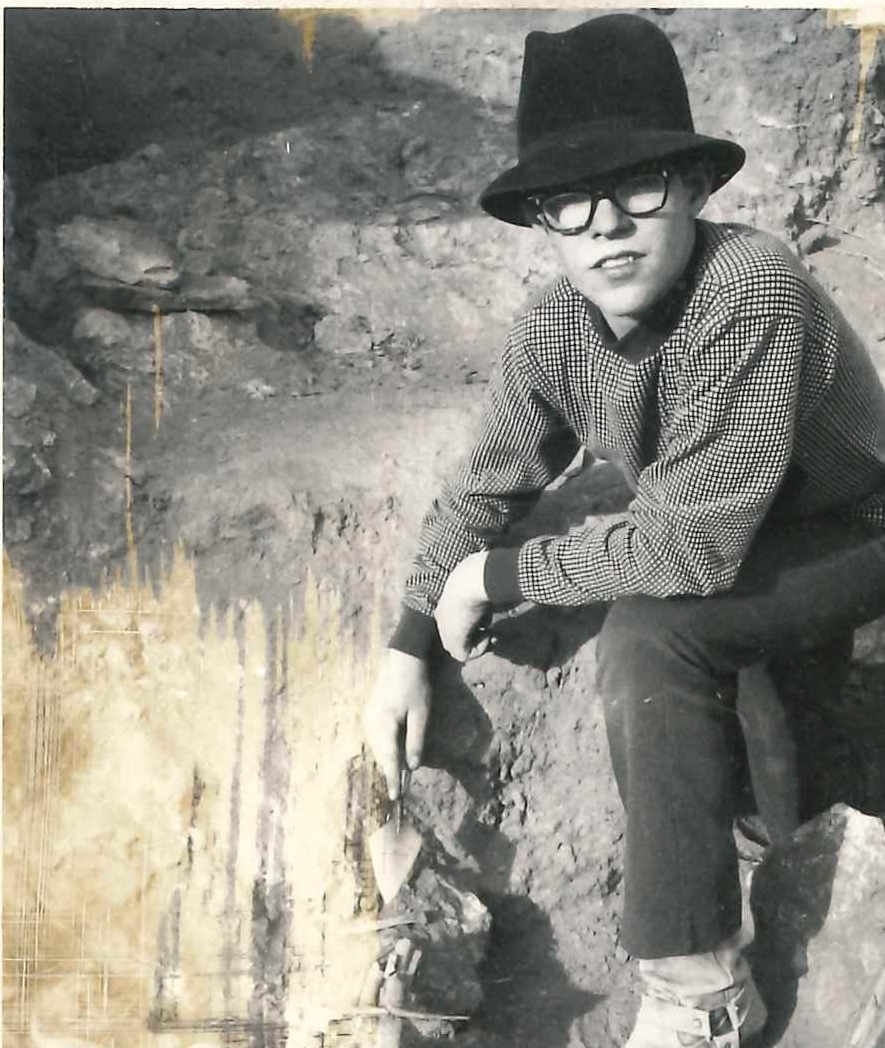 black and white photo of a boy excavating in dirt with a trowel, in a hat and glasses
