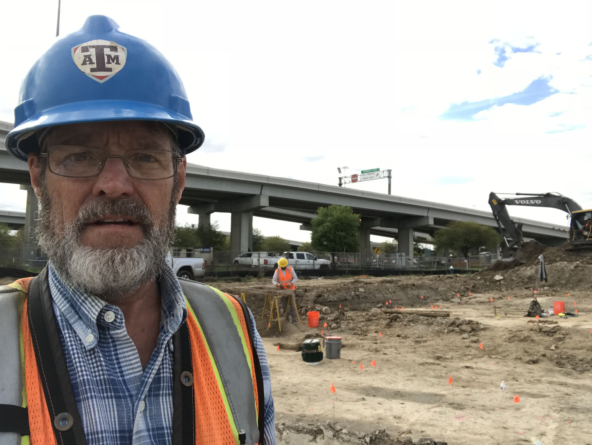 photo of a man standing near a highway overpass wearing a hard hat and orange vest, with an excavation behind him