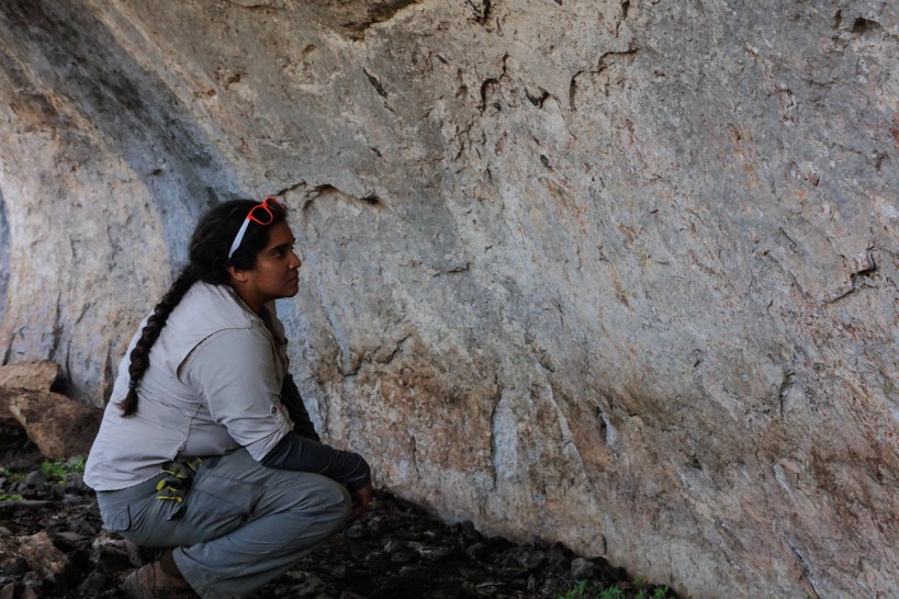 a woman kneeling in a rock shelter looking at faint pictographs