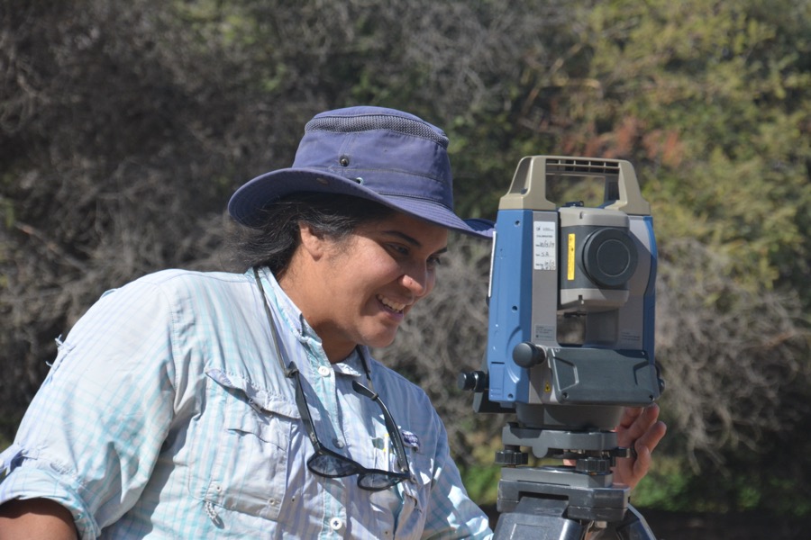 a woman wearing a sun hat operating a total data station with trees and brush behind her