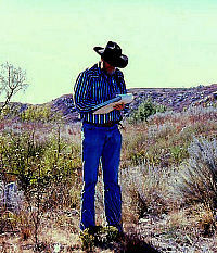 Photo of tall man wearing cowboy hat and standing in arid valley taking notes on a clipboard.