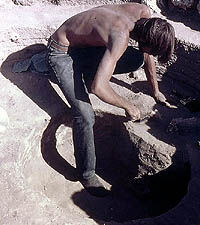 Photo of bare-shirted young man bending over and digging with a trowel.